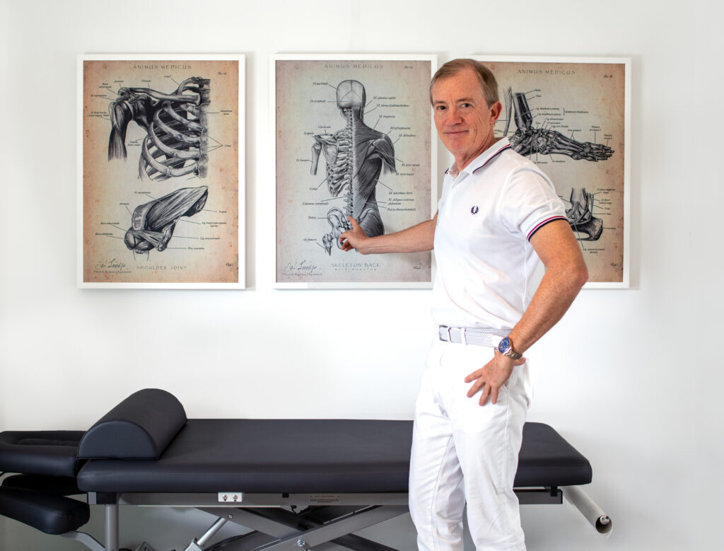 Dr. Christian Merettig im Behandlungsraum vor zwei anatomischen Bildtafeln